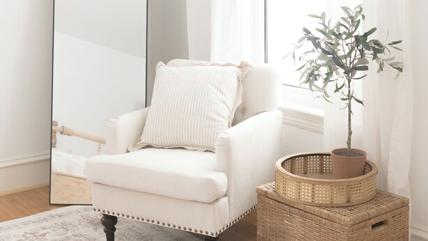 Ein weißer Sessel mit Kissen, ein großer Spiegel, ein Blumentopf, ein gewebter Korb und ein gemusterter Teppich.