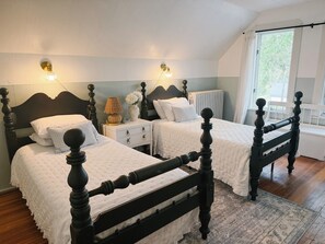 Ein Schlafzimmer mit zwei Betten, Nachttischlampen, einem Nachttisch, einem Heizkörper und einem Fenster mit Vorhängen.