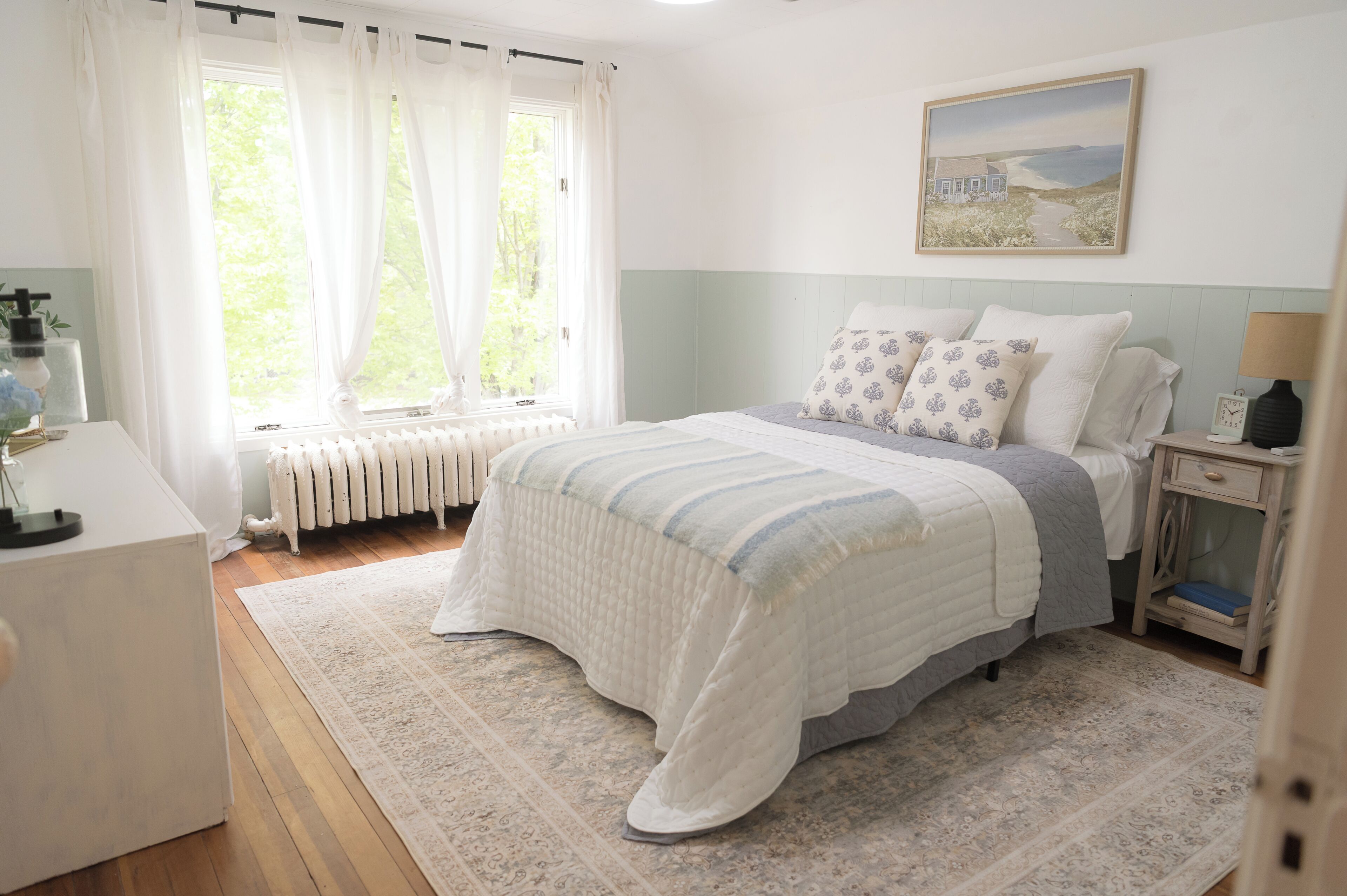 A bedroom with a bed, a nightstand, a window with curtains, a radiator, and a painting on the wall.