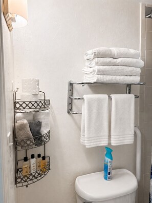 Ein Badezimmer mit Toilette, einem Handtuchhalter mit gefalteten Handtüchern, einem Metallregal mit Pflegeprodukten und einer Duschkabine.
