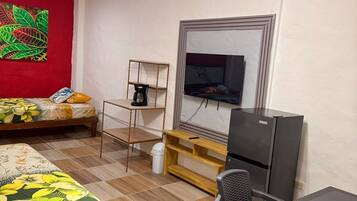 Una habitación con cama, escritorio, silla, televisor y estante con cafetera.