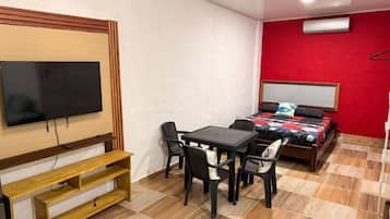 A room with a TV, a bed with a red wall, a table with chairs, and a wooden bench.