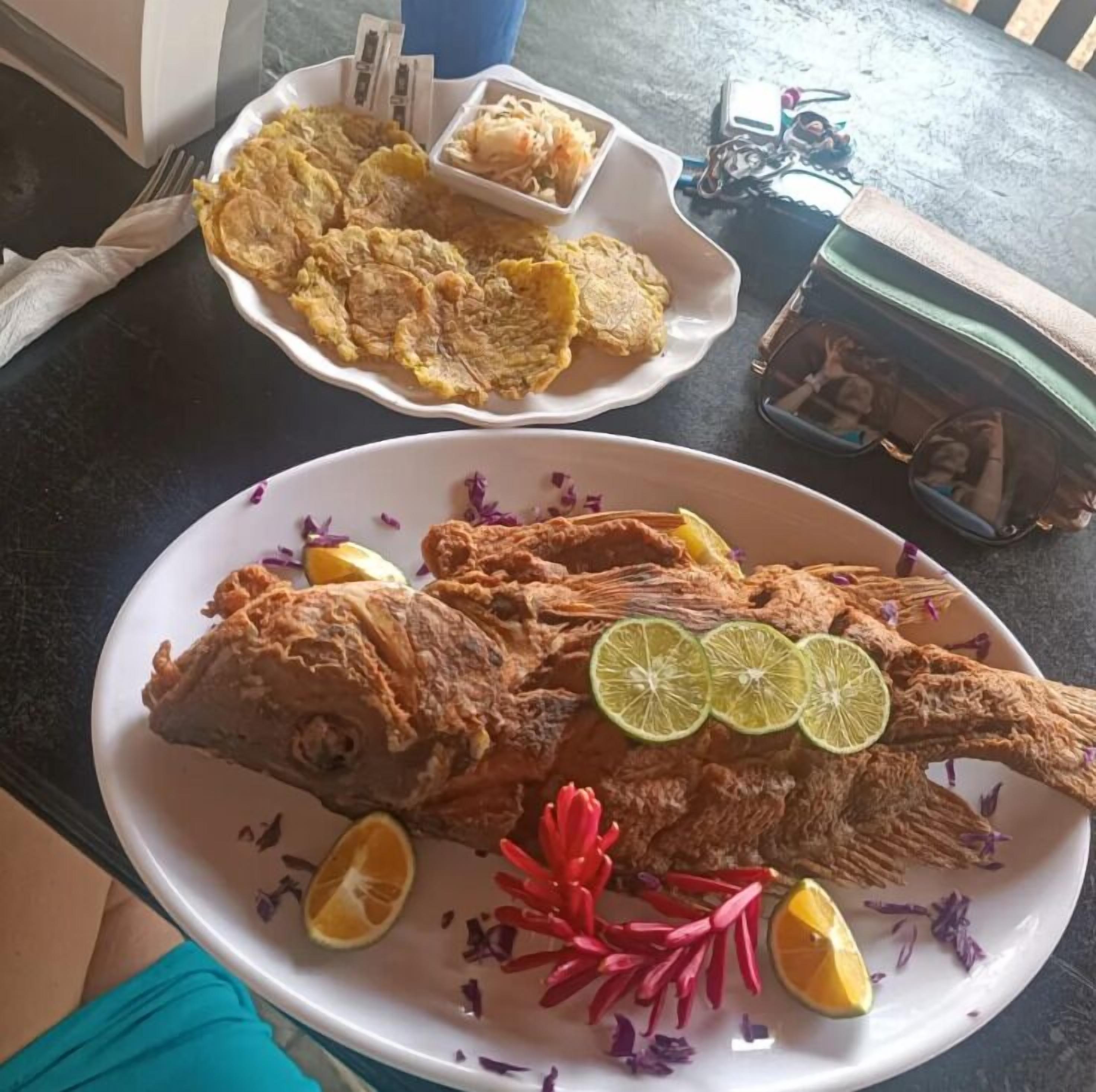 A dining table with plates of fried fish and plantains, a wallet, and a camera.