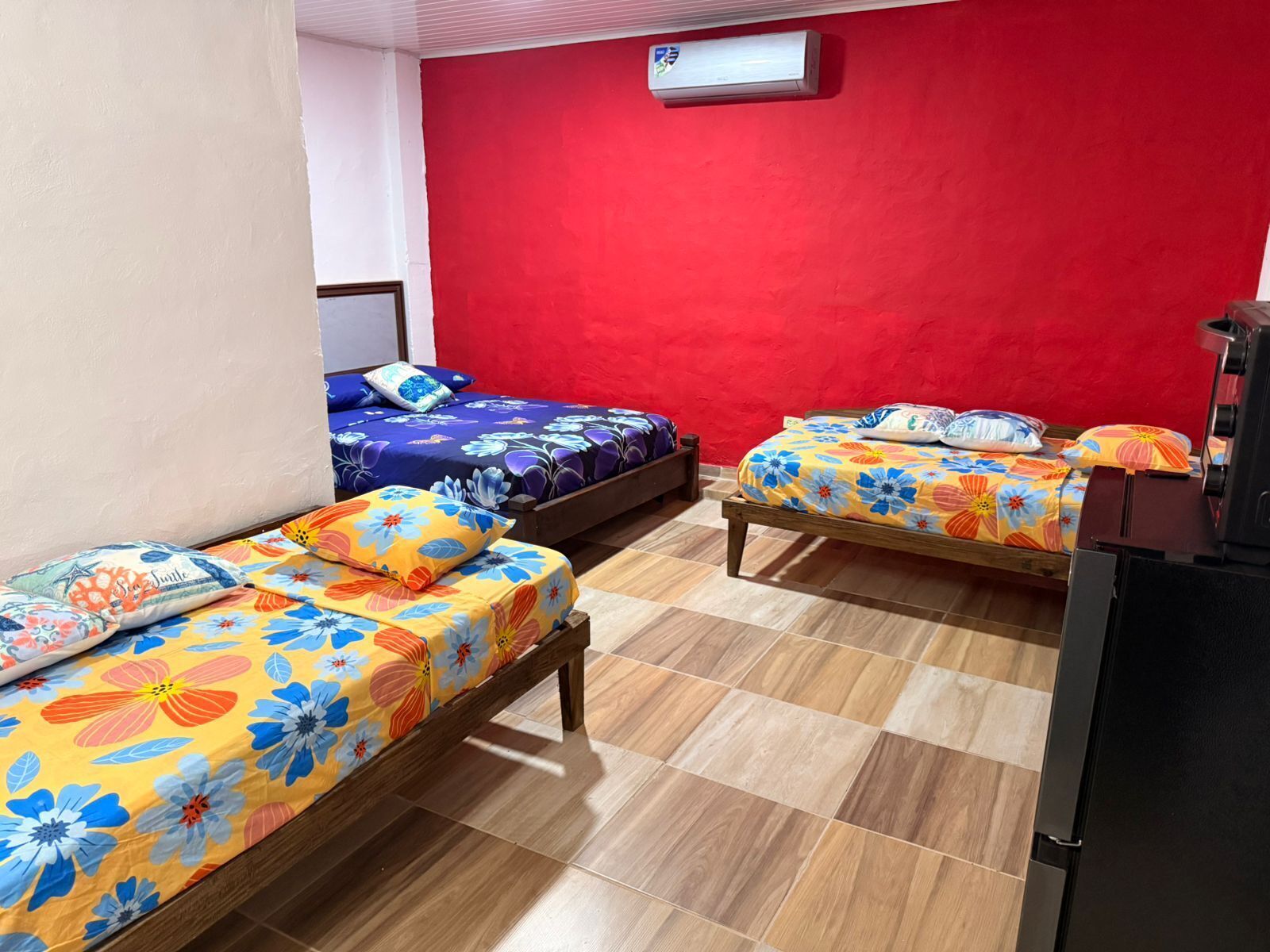 Una habitación con tres camas de madera, cada una con ropa de cama floral de colores, una pared roja y suelo de baldosas.