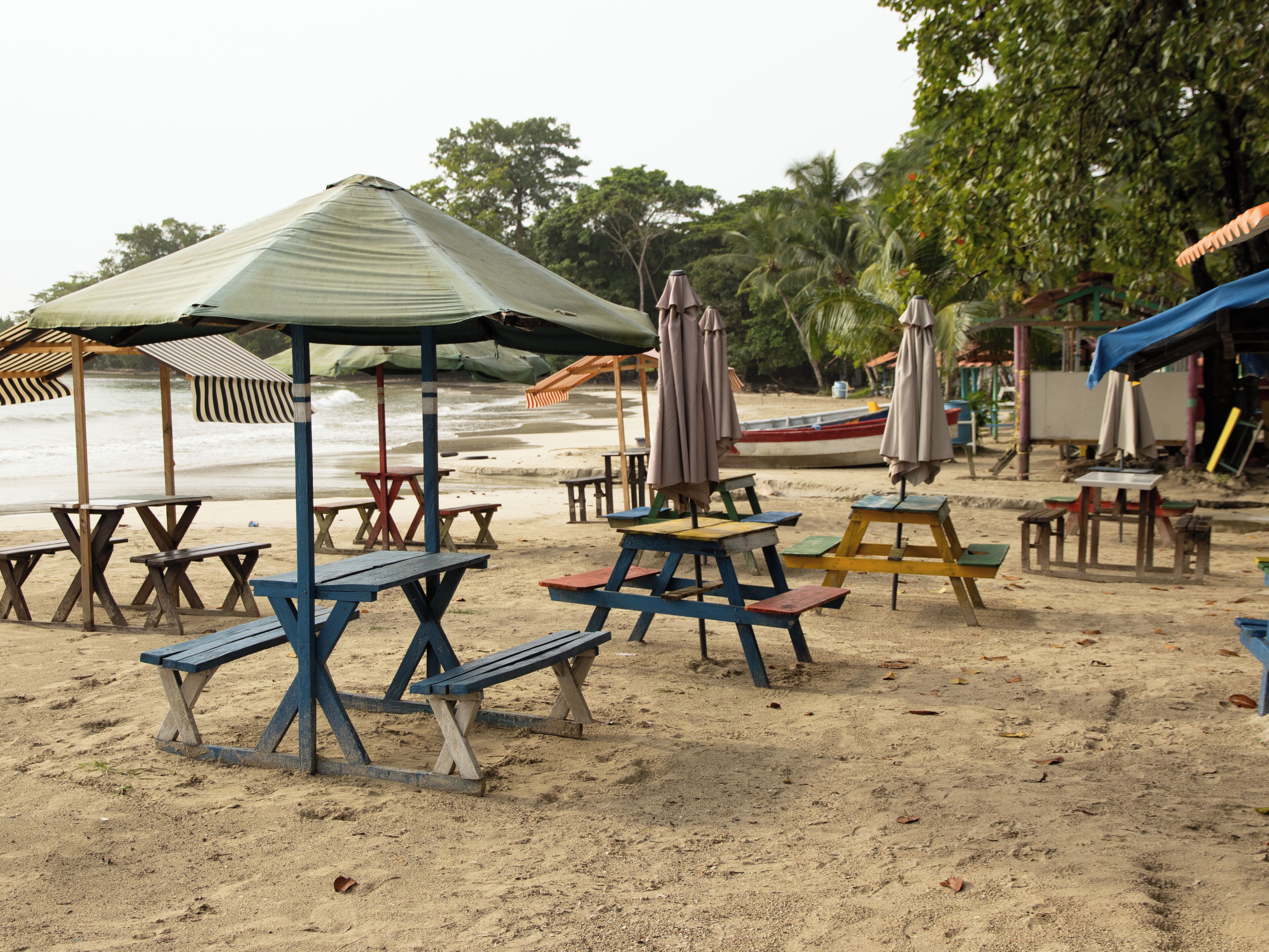 A beach with wooden picnic tables and umbrellas, a boat in the background, and trees.