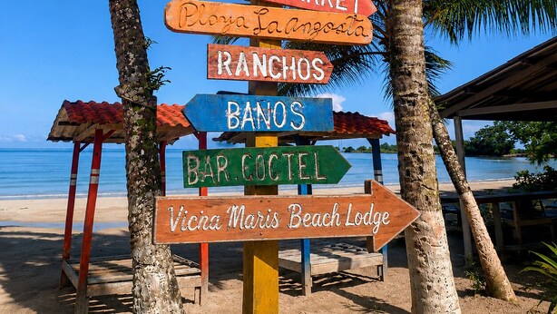A wooden signpost with multiple directional signs, including 'BIENVENIDOS,' 'BEACH MARKET,' 'RANCHOS,' 'BANOS,' 'BAR COCOTEL,' and 'Vicna Maris Beach Lodge.' - Vicina Maris Beach Lodge (Maria Chiquita)