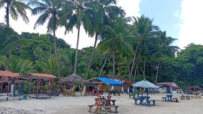 A beach with thatched-roof structures and tables, surrounded by palm trees. - Vicina Maris Beach Lodge (Maria Chiquita)