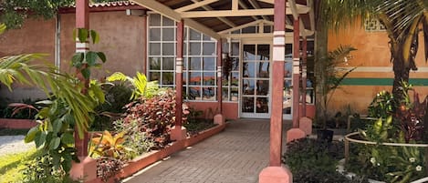 A covered walkway leading to a building with a glass entrance, surrounded by lush greenery and palm trees.