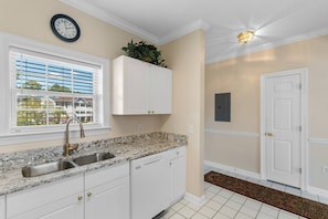 A kitchen with white cabinets, a granite countertop, a stainless steel sink, a clock, and a door with a light fixture above it. - Golf Course View | Spacious Coastal living (North Myrtle Beach)