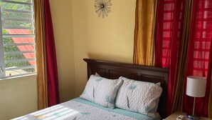 Una cama con colcha, almohadas, una mesita de noche con lámpara y un reloj de pared.