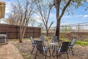 An outdoor seating area with metal chairs and a small table, surrounded by a wooden fence and trees. - Private Fenced Yard | Walk to AT&T + Globe Life! (Arlington)