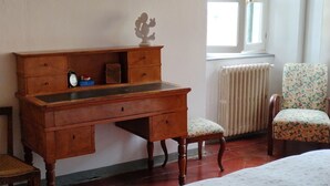 Een kamer met een houten bureau, een stoel, een bed met een bloemenprint en een radiator, plus een raam.