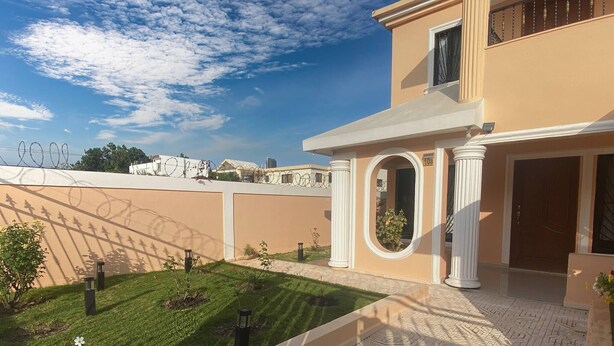 A building with a beige exterior, a paved courtyard with a patterned floor, and a small garden area with a flower. - Large House With AC Wifi Parking Ideal for Families (San Juan de la Maguana)