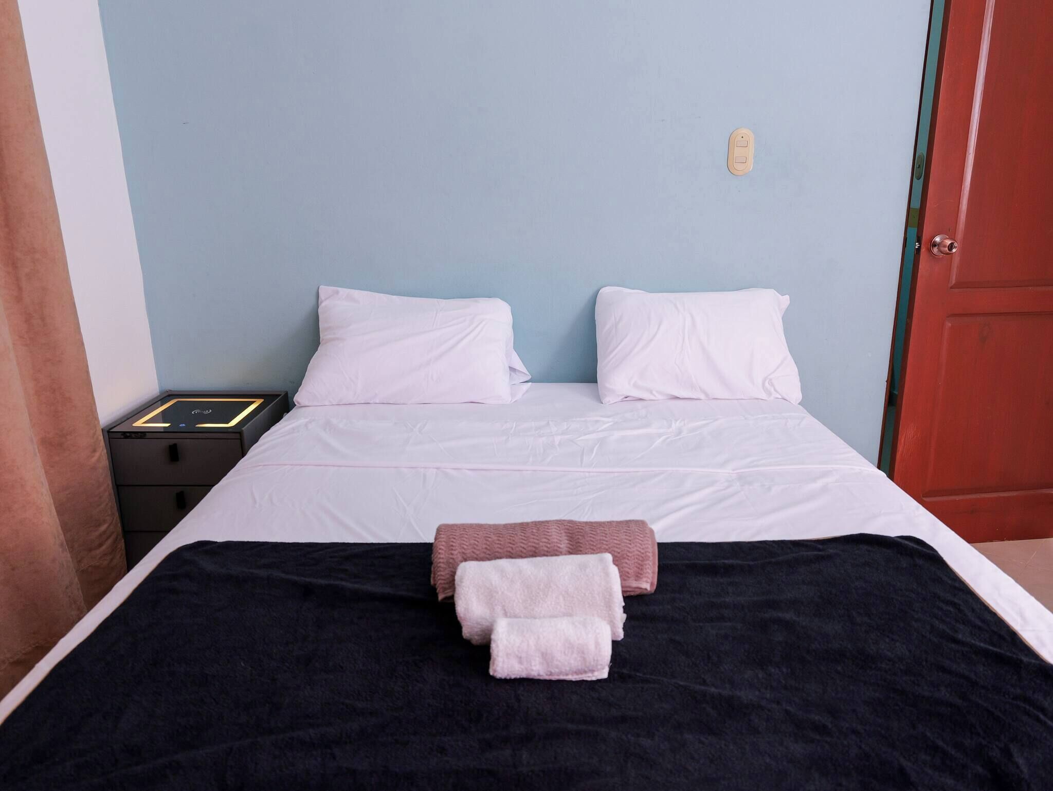 A neatly made bed with white linens, a black blanket, and two towels, a red door, and a light blue wall.