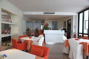 A hotel lobby with multiple dining tables, a reception desk, and a visible restroom sign. - Hotel San Carlo (Lignano Sabbiadoro)