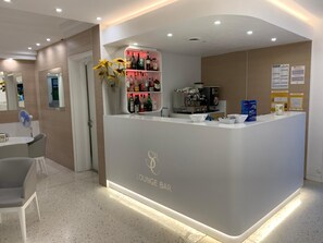 A lounge bar with a white counter, a fan, and a shelf with bottles. - Hotel San Carlo (Lignano Sabbiadoro)