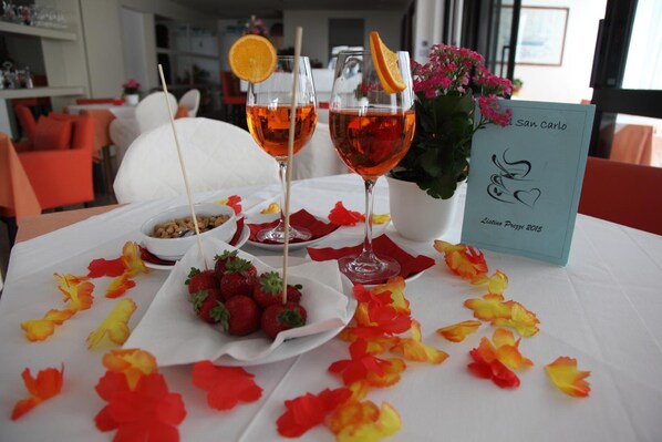 A table with two glasses of red beverage, a bowl of strawberries, and a flower arrangement. - Hotel San Carlo (Lignano Sabbiadoro)