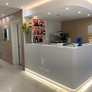 A lounge bar with a white counter, shelves with bottles, a coffee machine, and a vase with sunflowers. - Hotel San Carlo (Lignano Sabbiadoro)