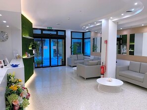 A modern lobby with a reception desk, seating area, and a large window. - Hotel San Carlo (Lignano Sabbiadoro)