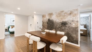 A dining area with a wooden table and chairs, a wall with a nature-themed mural, and a view into another room with a bed.