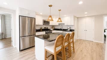 A modern kitchen with a stainless steel refrigerator, a microwave, and a built-in oven. There are two wooden chairs at the island counter, and pendant lights hang above.
