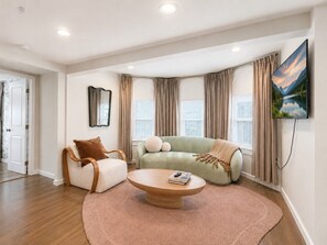 A modern living room with a sofa, armchair, and coffee table, a flat-screen TV mounted on the wall, and large windows with curtains. - Free Parking | Walk to Yale & Frank Pepe’s (New Haven)