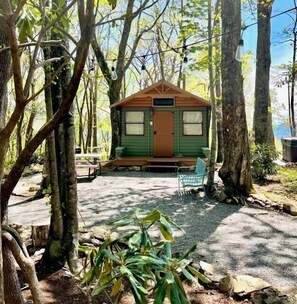 Een klein vakantiehuisje met een houten terras, omringd door bomen en een grindpad.