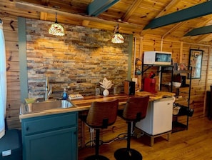 Cocina rústica con pared de piedra, techo de madera, un lavamanos, taburetes de bar, microondas y un refrigerador pequeño.