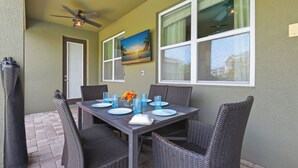 A dining area with a table set for four, wicker chairs, a ceiling fan, and a painting of a beach scene. - WOW! 6BR Pool Villa w/ Game Room Near Disney (Kissimmee)