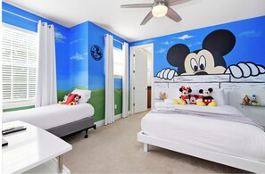 Ein Hotelzimmer mit zwei Betten, einem Deckenventilator, einem Fenster mit Jalousien und einem Wandgemälde mit Mickey Mouse.