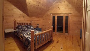 A wooden cabin bedroom with a bed, a chair, a nightstand, and a sliding glass door leading to a balcony.