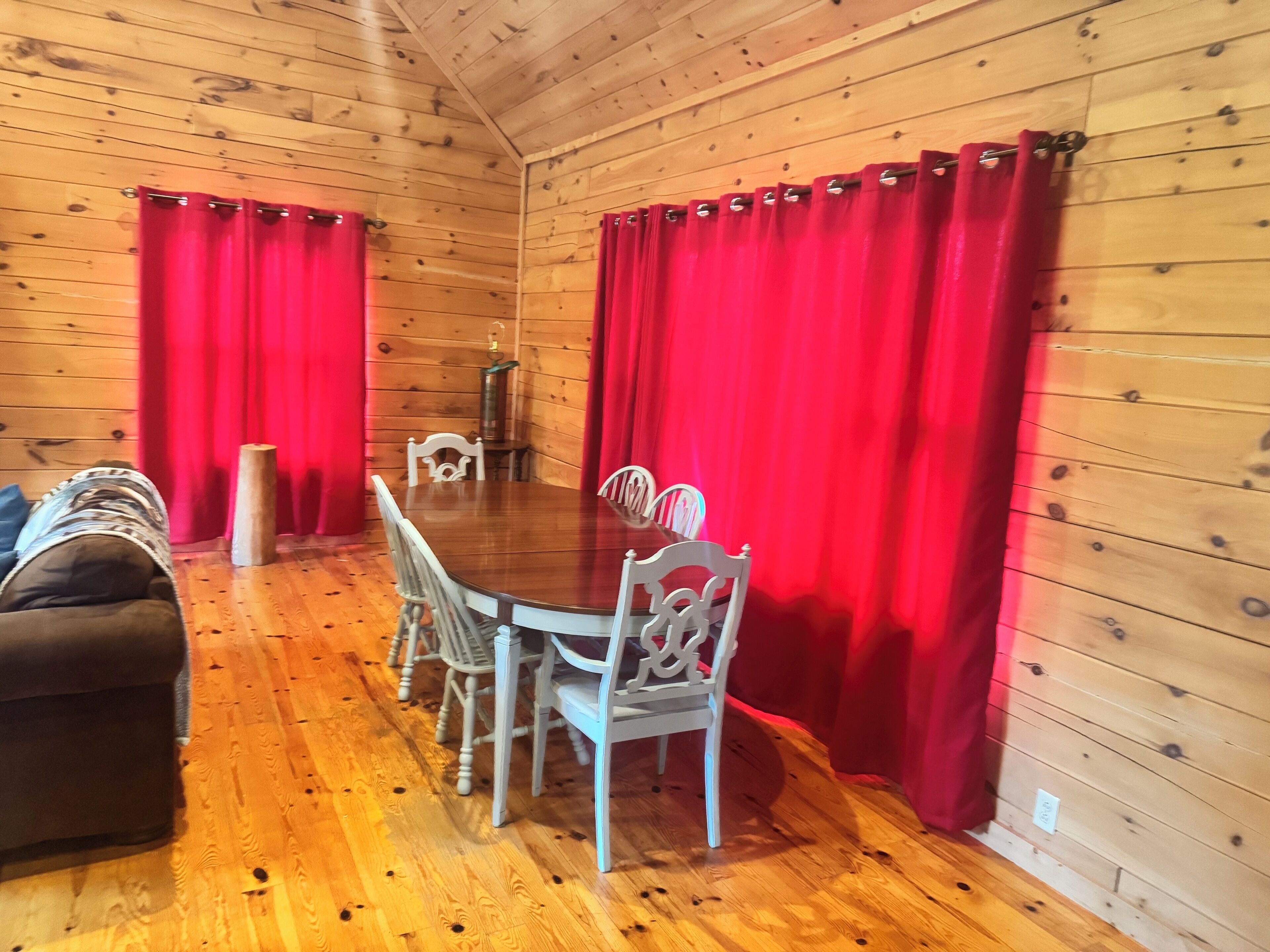 L’intérieur d’un chalet en bois comprend une table à manger, quatre chaises, un canapé et des rideaux rouges.