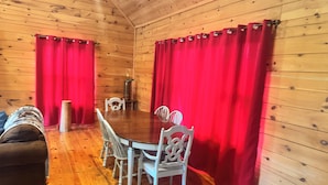 A wooden cabin interior with a dining table, four chairs, a sofa, and red curtains.