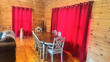 L’intérieur d’un chalet en bois comprend une table à manger, quatre chaises, un canapé et des rideaux rouges.