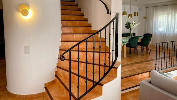Un escalier avec une rampe noire, un coin repas avec une table et des chaises, et un mur blanc agrémenté d’un luminaire.