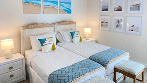 A double bed with a white and blue patterned bedspread, two nightstands with lamps, a ceiling fan, and wall art depicting beach scenes. - Beautiful 4Ba/Br, oceanfront, & ground floor- one of only several on the island! (South Padre Island)