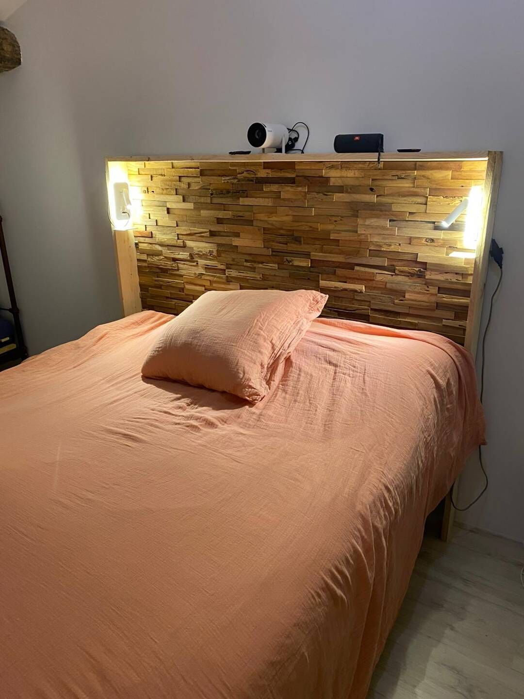 Quarto com cabeceira de madeira, cama com roupa de cama cor de pêssego e mesa de cabeceira com abajur.