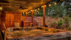 A hot tub with a wooden deck and string lights, surrounded by outdoor furniture and greenery. - Luxury Mountain House|HotTub|Decks|Shuffleboard (Mineral Bluff)