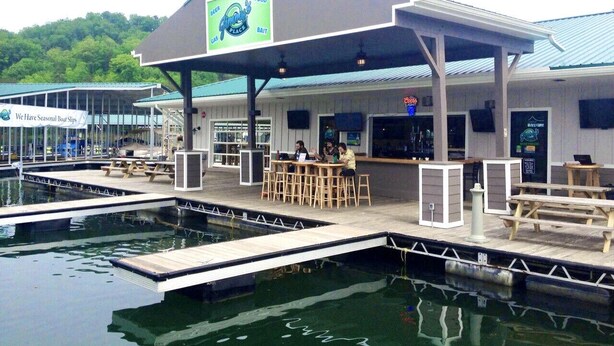 Un bar situé au bord de l’eau, doté d’un espace de repos couvert, d’un quai avec des bateaux et d’un panneau indiquant la possibilité de louer des bateaux pendant certaines périodes de l’année.