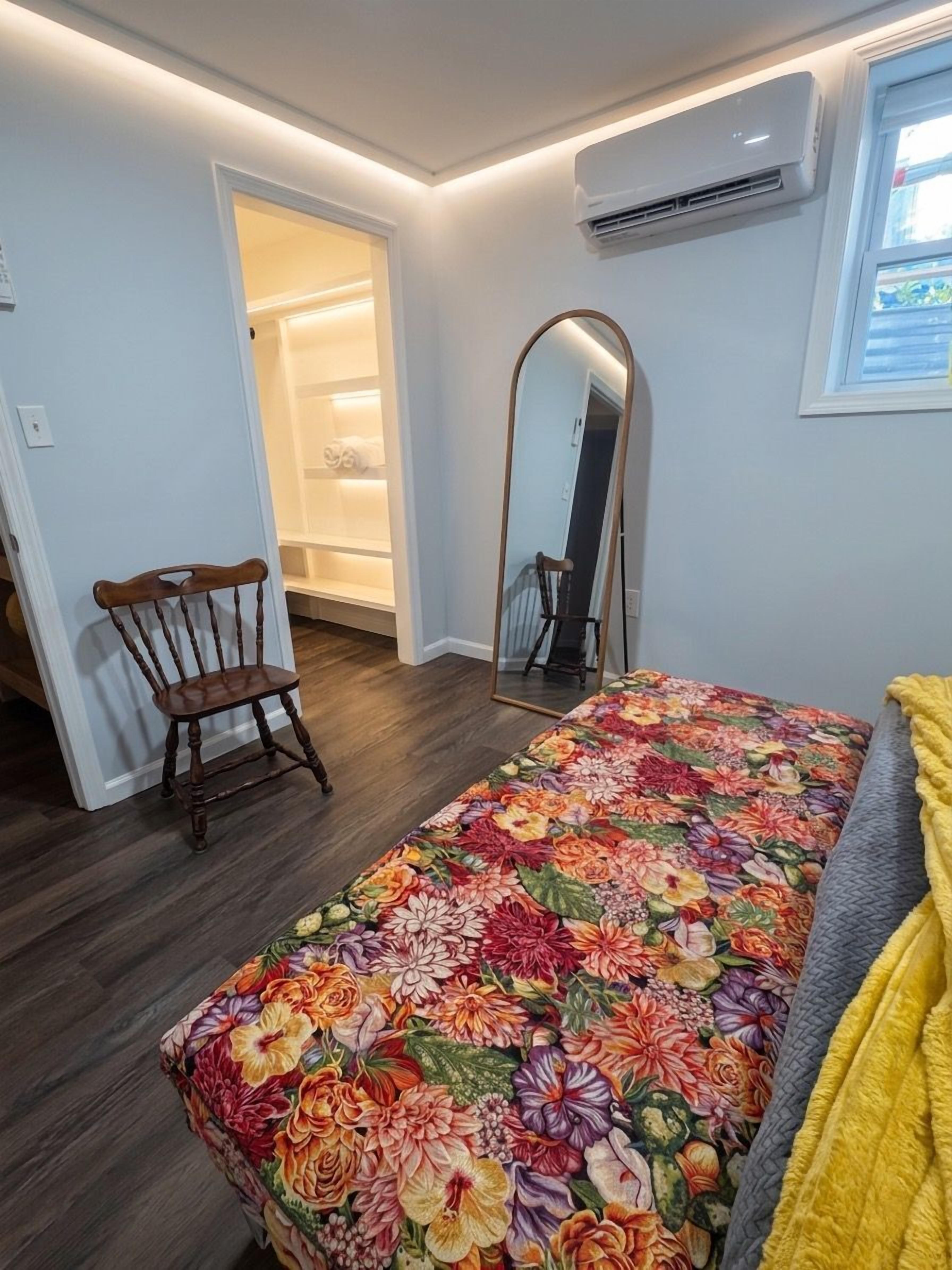 Een slaapkamer met een bloemenbedekking, een houten stoel, een spiegel en een airconditioning.