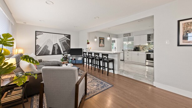 A modern living room with a sofa, a coffee table, and a TV. There is a dining area with a bar and chairs. The kitchen is visible in the background with white cabinets and stainless steel appliances. - Modern 4BR Retreat: Private Yard - 10 Min to DEN (Arvada)