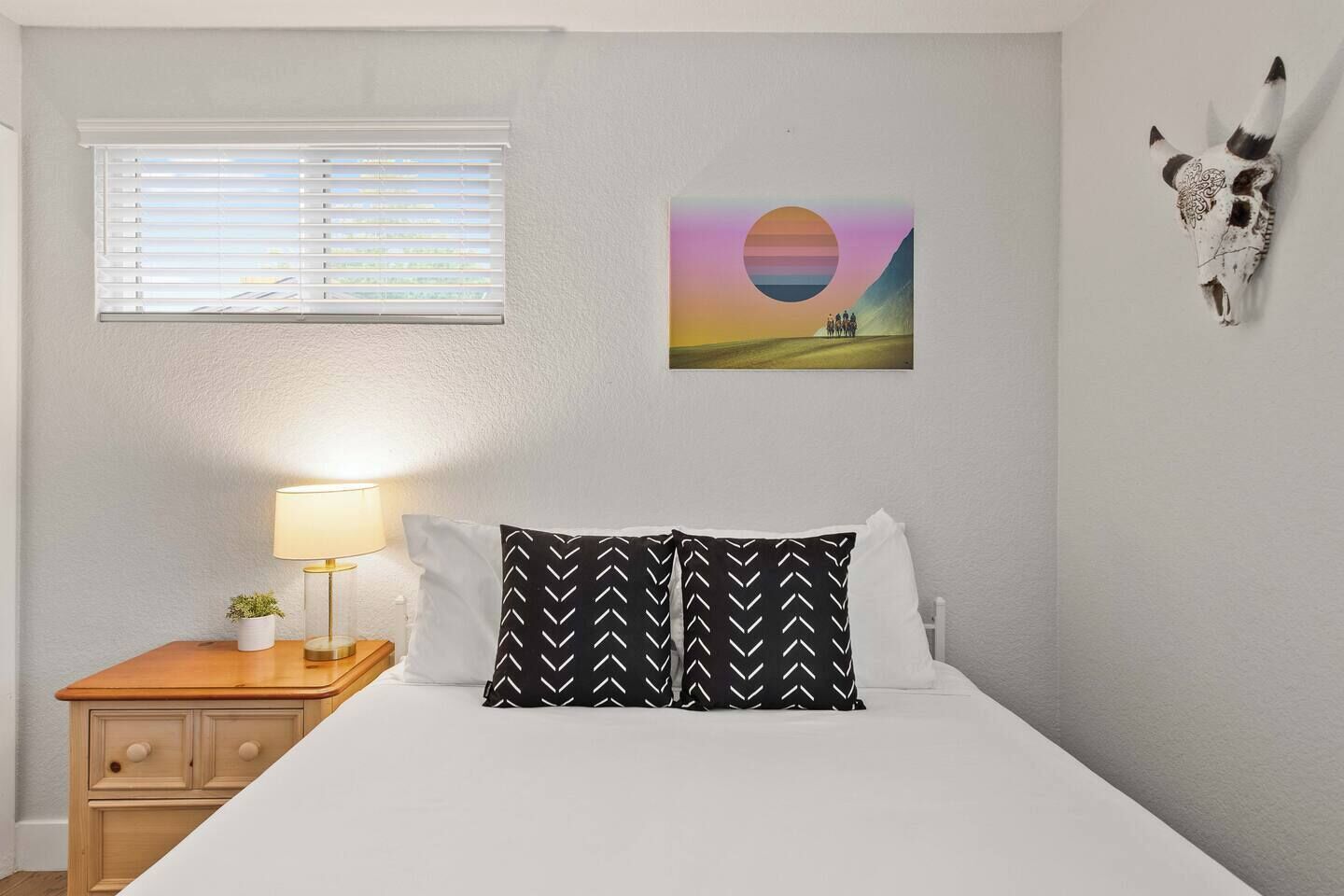 Un dormitorio con cama, una mesita de noche con lámpara, un cuadro colgado en la pared y una decoración de cráneo de vaca montado en la pared.
