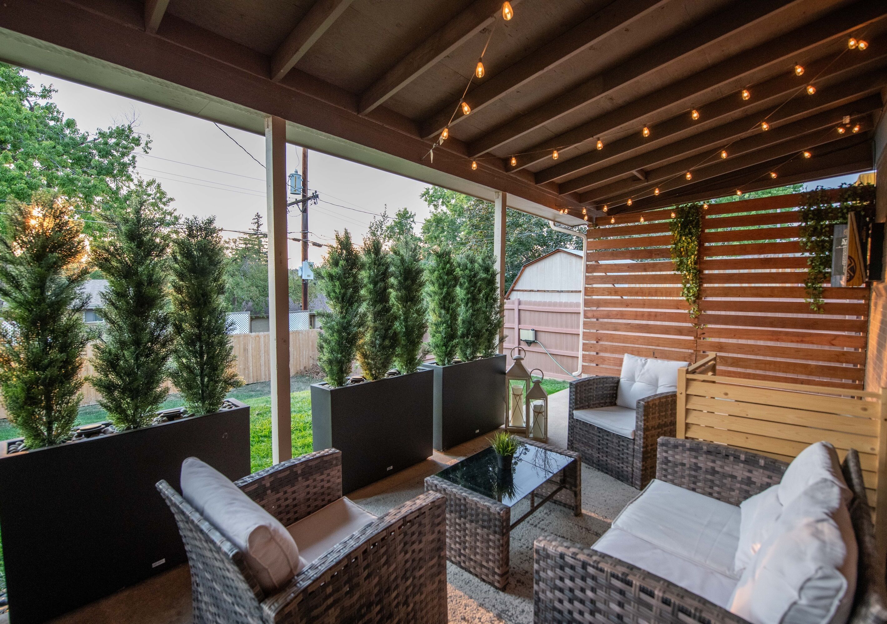Un patio exterior con muebles de mimbre, una mesa de vidrio y una valla de madera con plantas.
