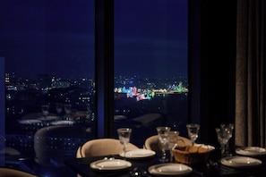 Une table dressée pour le dîner, avec une vue sur la ville à travers de grandes fenêtres.