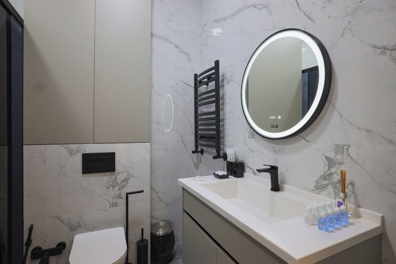 Une salle de bain moderne avec un mur en marbre blanc, un grand miroir rond doté d’un éclairage à LED, un porte-serviettes noir, un lavabo blanc avec un robinet noir et une poubelle.