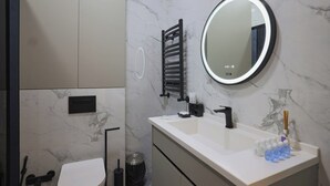 Ein modernes Badezimmer mit weißer Marmorwand, einem runden Spiegel mit LED-Beleuchtung, einem schwarzen Handtuchhalter, einem weißen Waschbecken mit schwarzem Wasserhahn und einem Mülleimer.