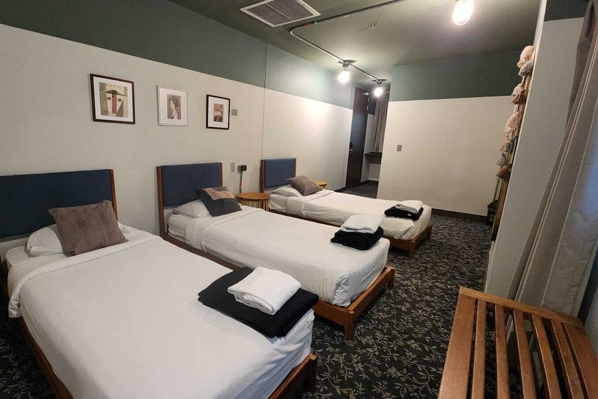 A hotel room with three single beds, a wooden bench, a round wooden stool, and a wall-mounted heater.