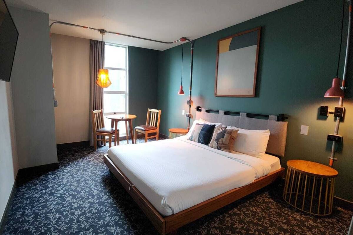 A hotel room with a bed, a desk, a chair, a small table, and a TV mounted on the wall.