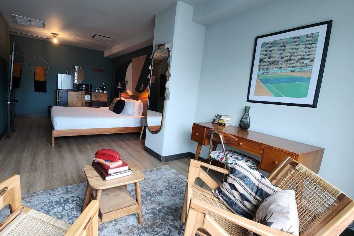 A hotel room with a bed, a desk, a chair, a small table with books, a mirror, and a wall-mounted TV.