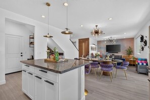 A modern kitchen with a central island, pendant lighting, and a dining area with purple chairs. - Wanderlust | Pool+Spa, Sunken BBQ, FirePit, Games! (Coachella)